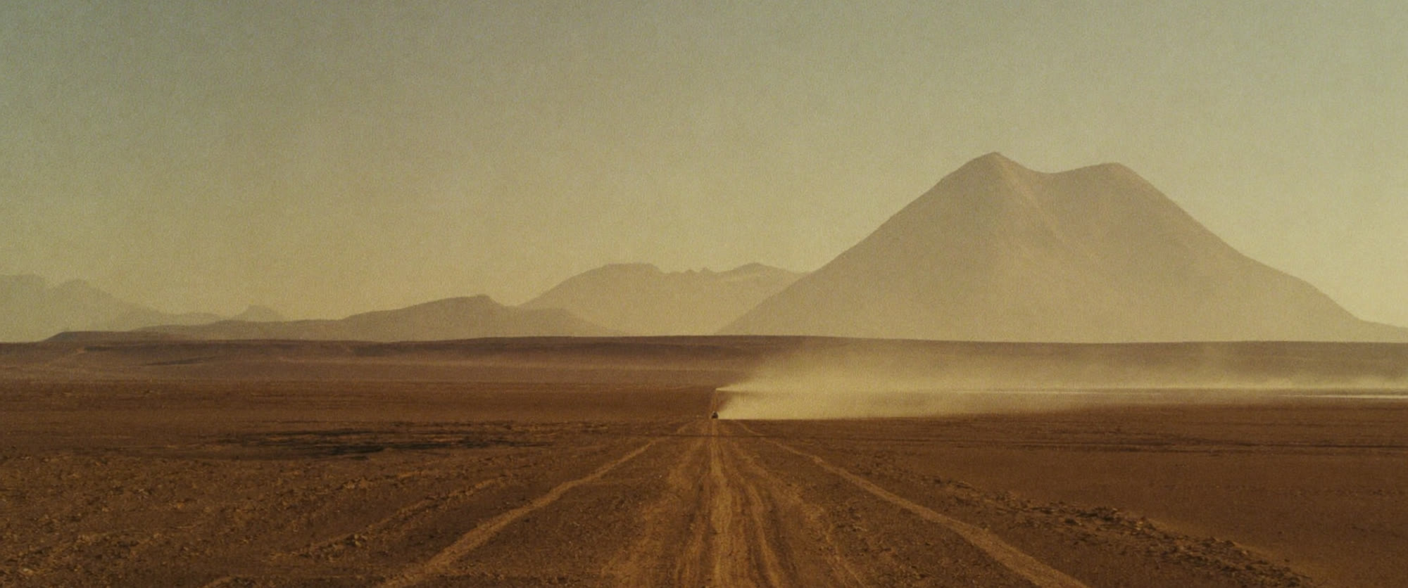 The approach road seen from a rise, a vehicle far ahead trailing dust across the plain.