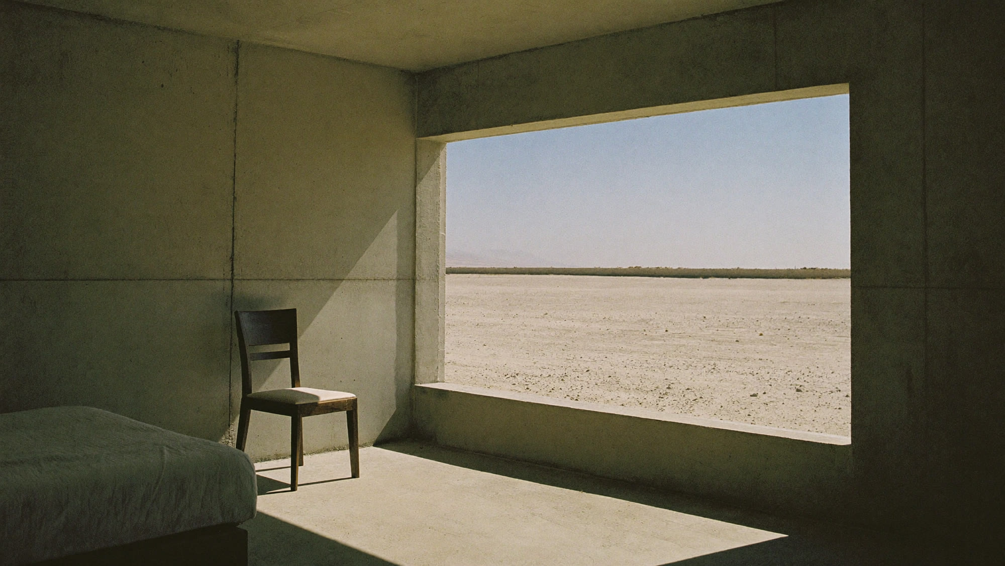 A bare concrete room with a bed, a wooden chair, and one enormous window looking out onto flat pale desert.