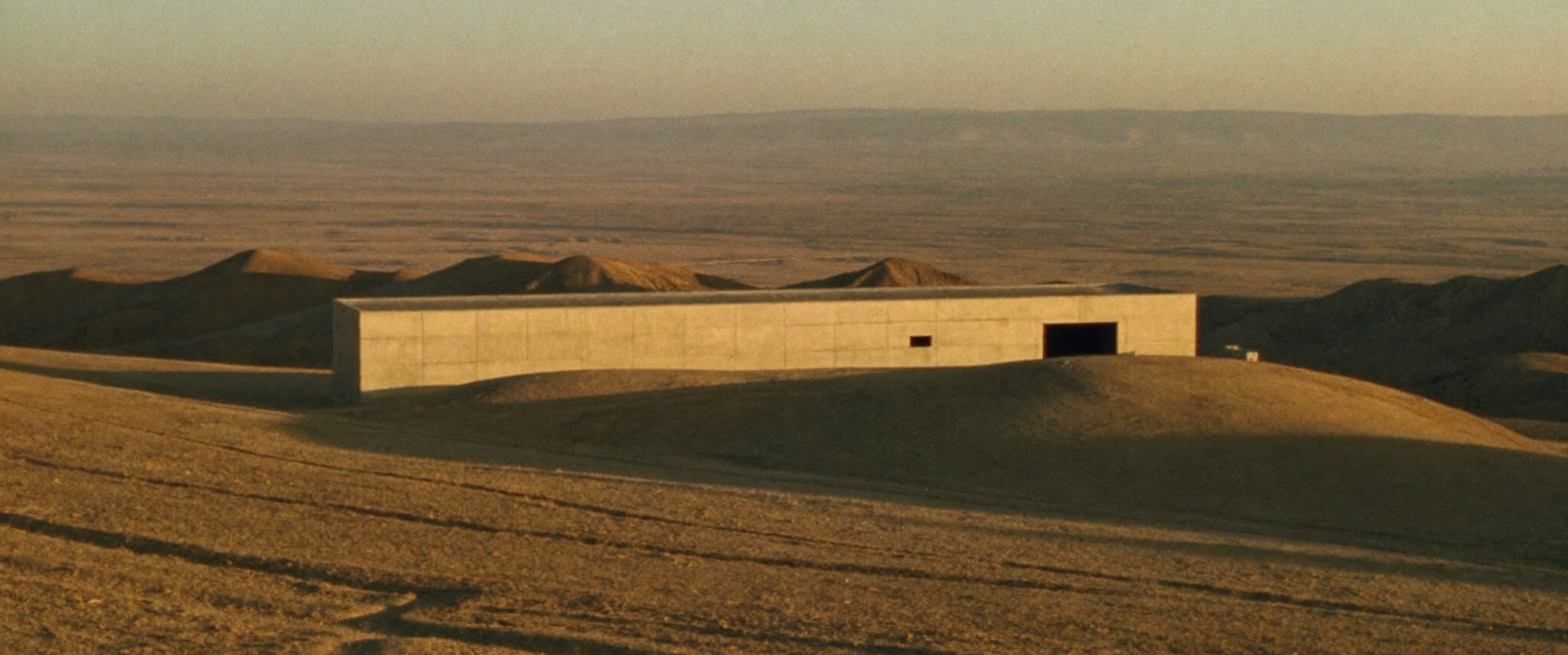 A long, low board-formed concrete building set into a ridge above an enormous pale desert basin at low sun.